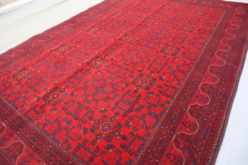 Op de afbeelding: Een rood tapijt met een geometrisch patroon in blauw en goud. Het tapijt heeft een rand met een herhalend ontwerp.