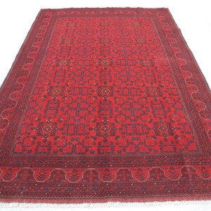 Op de afbeelding: Een rood tapijt met een gedetailleerd geometrisch patroon in tinten blauw en cr&egrave;me. Het tapijt heeft een franje rand.