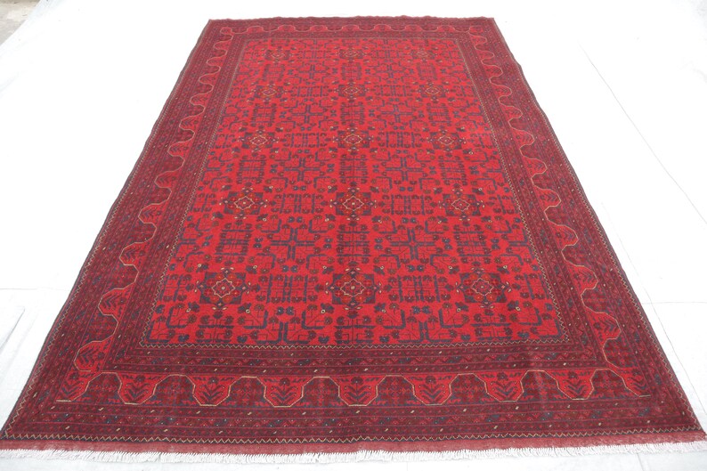 Op de afbeelding: Een rood tapijt met een gedetailleerd geometrisch patroon in tinten blauw en cr&egrave;me. Het tapijt heeft een franje rand.