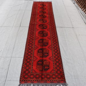 May include: A long, rectangular red rug with a repeating geometric pattern in black. The rug has a fringe on both ends and is likely made of wool. The design features a central series of black medallions.