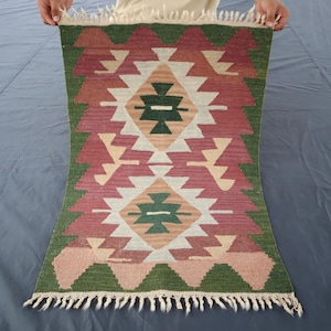 May include: A traditional Turkish rug with a geometric pattern in shades of red, green, and white. The rug has a fringe border.