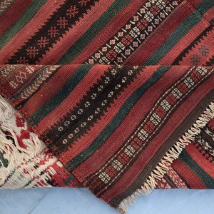 May include: A close-up of a red, green, and black striped rug with a fringe border. The rug is folded in half, showing the intricate woven pattern.