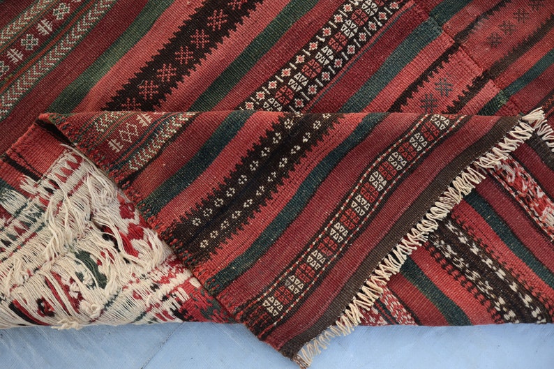 May include: A close-up of a red, green, and black striped rug with a fringe border. The rug is folded in half, showing the intricate woven pattern.