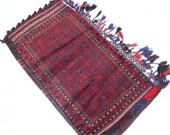 May include: A red, black, and blue patterned rug with fringe on all sides. The rug has a geometric design and is made of wool.