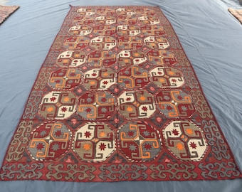 May include: A rectangular, patterned rug with a red, cream, gray, and orange geometric design. The rug features repeating circular patterns with star-like shapes and a decorative border. The rug is laid out on a gray surface.