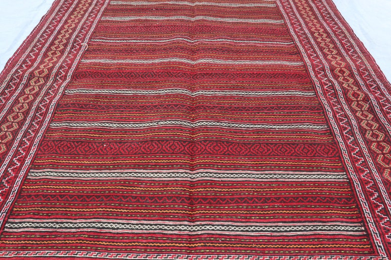 Puede incluir: Una alfombra roja, blanca y negra a rayas con intrincados patrones geom&eacute;tricos en el borde. La alfombra tiene un aspecto y una sensaci&oacute;n tradicionales tejidos.