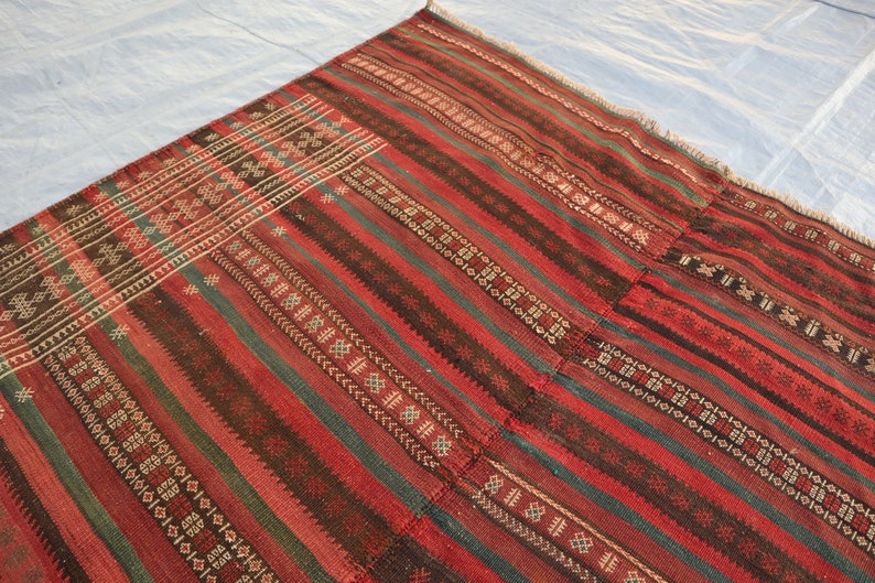 May include: A red, green, and black striped rug with intricate geometric patterns woven into the fabric. The rug has a frayed edge and appears to be handmade.