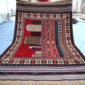 Op de afbeelding: Een rechthoekig, handgeweven tapijt met een rood middenveld en ingewikkelde geometrische patronen. De rand heeft een herhalend ontwerp in crème, bruin en bordeauxrood. Het tapijt heeft een gefranjerde rand en wordt getoond op een lichtblauwe ondergrond.