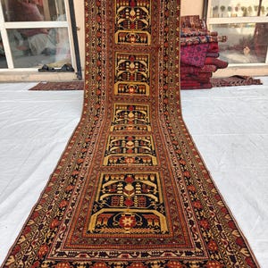 Puede incluir: Una alfombra pasillera larga y rectangular con un patrón repetitivo de motivos florales geométricos en azul oscuro, rojo y dorado. La alfombra tiene un borde ancho e intrincado con formas triangulares y florales, rematado con flecos cortos.