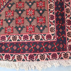 Puede incluir: Primer plano de una secci&oacute;n de una alfombra tejida a mano. La alfombra presenta un patr&oacute;n floral repetitivo en tonos rojos, verdes y marrones. Un borde azul oscuro con dise&ntilde;os florales y geom&eacute;tricos enmarca la alfombra, con una franja color crema.