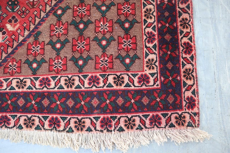 Puede incluir: Primer plano de una secci&oacute;n de una alfombra tejida a mano. La alfombra presenta un patr&oacute;n floral repetitivo en tonos rojos, verdes y marrones. Un borde azul oscuro con dise&ntilde;os florales y geom&eacute;tricos enmarca la alfombra, con una franja color crema.