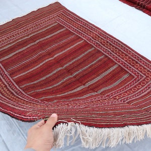 Puede incluir: Una alfombra tejida roja y blanca con un patr&oacute;n geom&eacute;trico y flecos. La alfombra est&aacute; doblada por la mitad y sostenida por una mano.