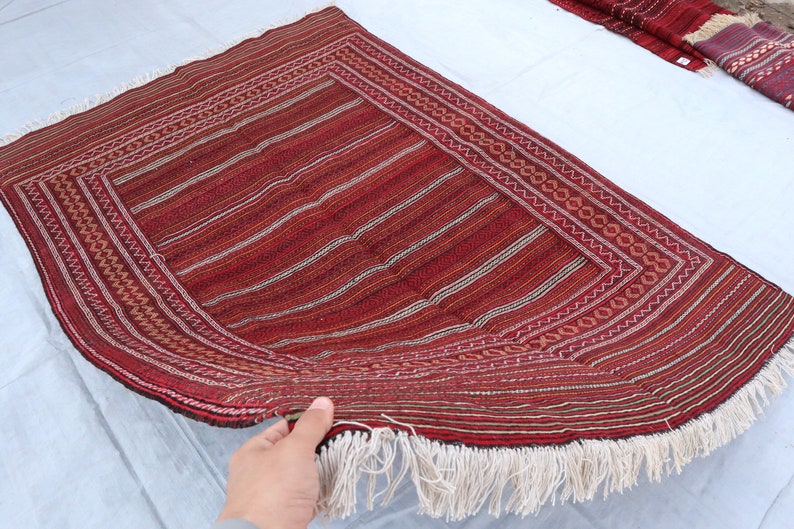 Puede incluir: Una alfombra tejida roja y blanca con un patr&oacute;n geom&eacute;trico y flecos. La alfombra est&aacute; doblada por la mitad y sostenida por una mano.