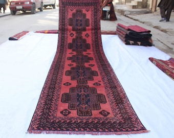 Puede incluir: Una alfombra pasillera larga y rectangular con un fondo rojo coral vibrante y una serie repetida de medallones geométricos en azul marino oscuro y negro. El borde muestra patrones angulares intrincados en tonos oscuros a juego.
