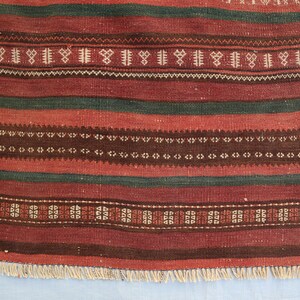May include: A red, green, and brown striped rug with a geometric pattern. The rug has a fringe border.