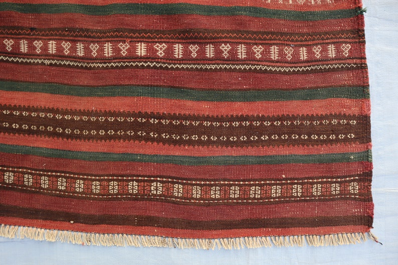 May include: A red, green, and brown striped rug with a geometric pattern. The rug has a fringe border.