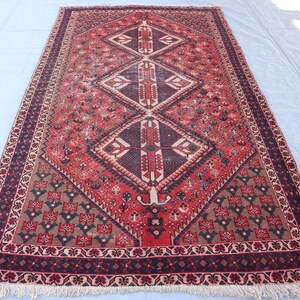 Puede incluir: Alfombra rectangular tejida a mano con un patr&oacute;n central de diamantes en tonos rojos, marrones y crema. Presenta intrincados dise&ntilde;os florales y geom&eacute;tricos, con un borde oscuro y bordes con flecos. Dimensiones aproximadas: 183 x 274 cm.