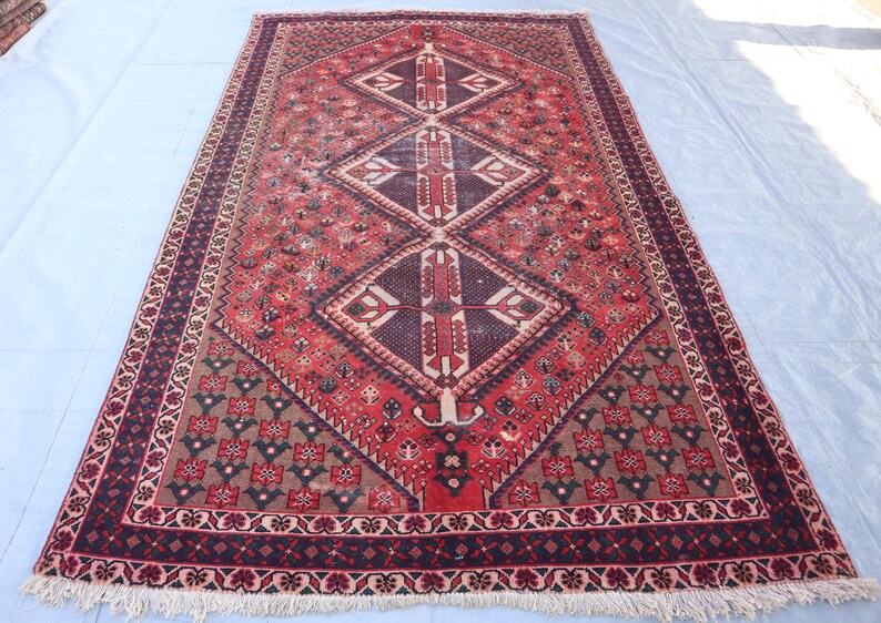 Puede incluir: Alfombra rectangular tejida a mano con un patr&oacute;n central de diamantes en tonos rojos, marrones y crema. Presenta intrincados dise&ntilde;os florales y geom&eacute;tricos, con un borde oscuro y bordes con flecos. Dimensiones aproximadas: 183 x 274 cm.