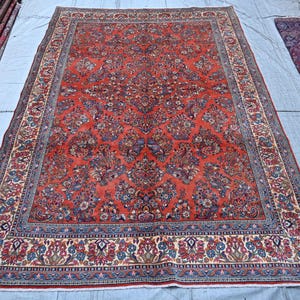 Puede incluir: Una alfombra rectangular con un campo rojo vibrante densamente cubierto de intrincados patrones florales y de enredaderas en tonos azul, crema y rojo oscuro. La alfombra tiene un borde ancho y detallado con motivos botánicos repetitivos.