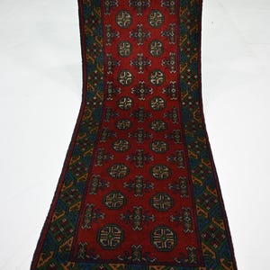 May include: A long, rectangular runner rug featuring a deep red background with repeating geometric octagonal motifs in blue, green, and cream. The border displays a complex diamond pattern in similar colors, finished with white fringe at the ends.