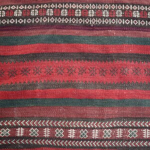 May include: A close-up of a red, black, and green woven rug with intricate geometric patterns. The rug has a traditional design and is made of natural fibers.