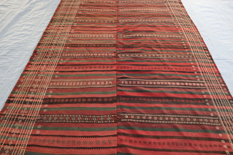 May include: A red, green, and black striped rug with intricate woven patterns. The rug has a fringed edge and is made of natural fibers.