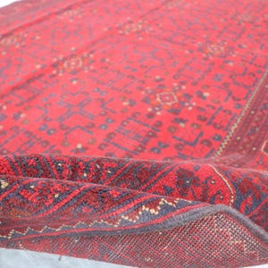Op de afbeelding: Een rood en blauw patroon tapijt met een franje rand. Het tapijt is dubbelgevouwen, waardoor de achterkant van het tapijt zichtbaar is.