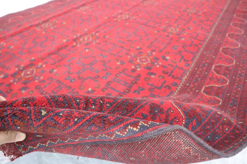 Op de afbeelding: Een rood en blauw patroon tapijt met een franje rand. Het tapijt is dubbelgevouwen, waardoor de achterkant van het tapijt zichtbaar is.
