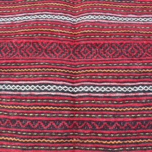 Puede incluir: Primer plano de una alfombra tejida roja, negra, blanca y naranja con un patr&oacute;n geom&eacute;trico repetitivo.