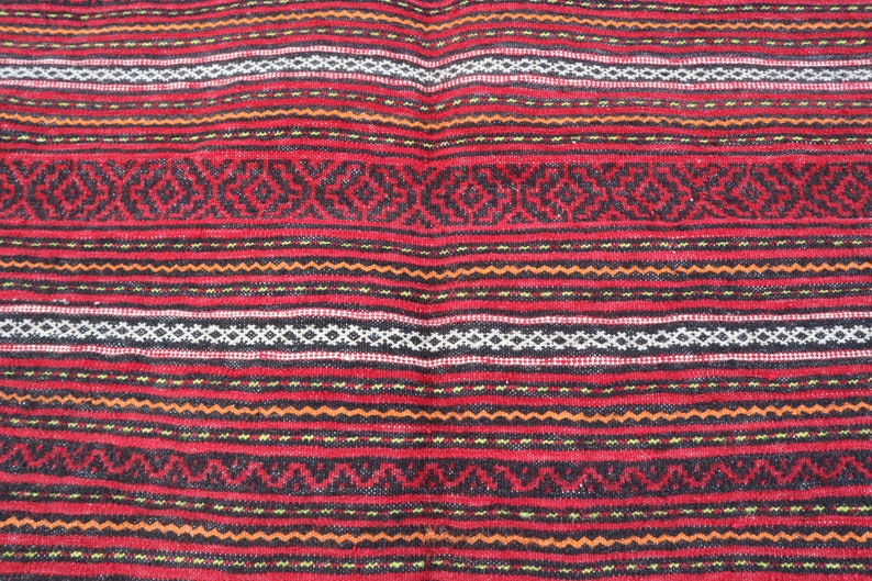 Puede incluir: Primer plano de una alfombra tejida roja, negra, blanca y naranja con un patr&oacute;n geom&eacute;trico repetitivo.