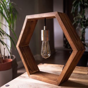 Puede incluir: Una base de lámpara hexagonal de madera con una bombilla colgante. La lámpara está hecha de madera marrón oscura y tiene un diseño simple y moderno.