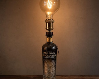 May include: A table lamp constructed from a clear glass Mermaid Spiced Rum bottle with a textured base. The bottle is topped with a brass-colored metal socket and a large, round, glowing Edison-style light bulb with a visible glowing filament inside.