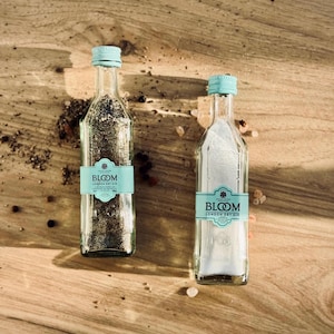 Può includere: Due piccole bottiglie di vetro con tappi blu e etichette che recitano "Bloom London Dry Gin". Le bottiglie sono su una superficie di legno con grani di pepe nero e sale rosa dell'Himalaya sparsi intorno.