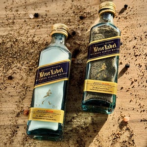 May include: Two small glass bottles filled with black peppercorns. The bottles are labeled with the Johnnie Walker Blue Label logo and text. The bottles are sitting on a wooden surface with scattered peppercorns.