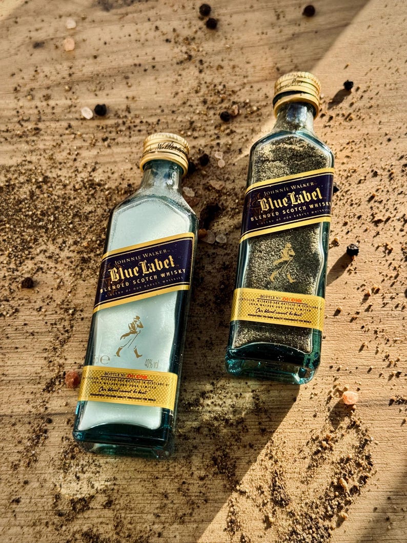 May include: Two small glass bottles filled with black peppercorns. The bottles are labeled with the Johnnie Walker Blue Label logo and text. The bottles are sitting on a wooden surface with scattered peppercorns.