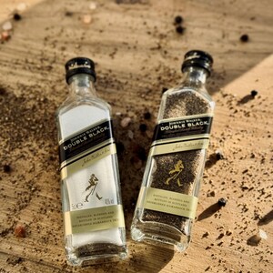 May include: Two small glass bottles filled with black peppercorns and white salt. Both bottles have a black label with gold lettering that reads "Johnnie Walker Double Black" and "40% vol". The bottles are sitting on a wooden surface with scattered black peppercorns and pink salt.