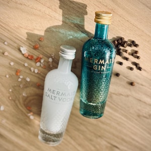 Peut inclure: Deux petites bouteilles d'alcool, l'une transparente et l'autre bleue, avec des lettres dorées. La bouteille transparente est étiquetée "Mermaid Salt Vodka" et la bouteille bleue est étiquetée "Mermaid Gin".
