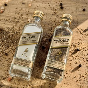 May include: Two clear glass bottles filled with salt and pepper, both labeled with the Johnnie Walker Gold Label Reserve logo. The bottles are sitting on a wooden surface with scattered salt and peppercorns.