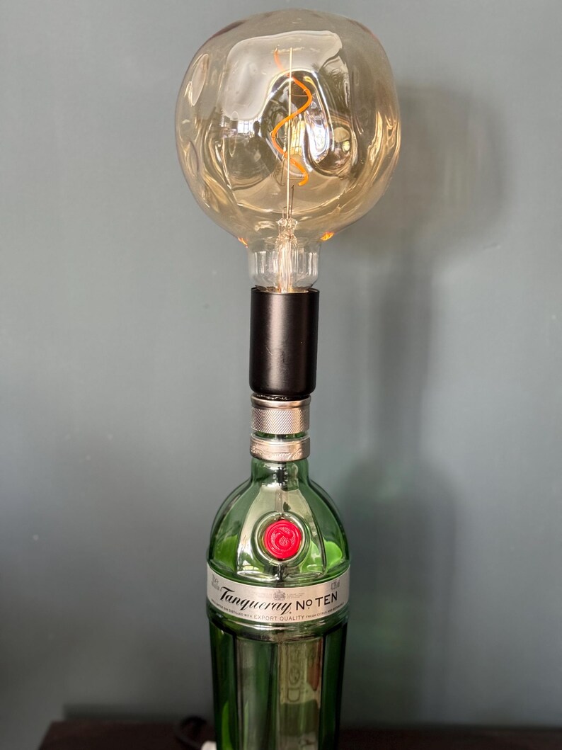 May include: A green glass bottle with a red cap and the text "Tanqueray No. TEN" is used as a lamp base. A large, round, clear glass light bulb with a coiled filament is attached to the top of the bottle.
