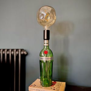 May include: A green glass bottle with a red label that says "Tanqueray No Ten" is used as a lamp base. A round, clear glass light bulb with a filament is attached to the top of the bottle. The lamp is sitting on top of two books, one with the title "Sapiens" and the other with the title "The Handmaid's Tale" by Margaret Atwood.