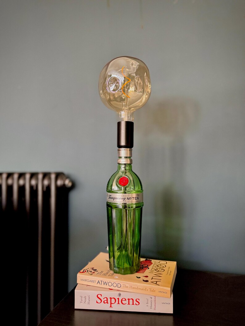 May include: A green glass bottle with a red label that says "Tanqueray No Ten" is used as a lamp base. A round, clear glass light bulb with a filament is attached to the top of the bottle. The lamp is sitting on top of two books, one with the title "Sapiens" and the other with the title "The Handmaid's Tale" by Margaret Atwood.