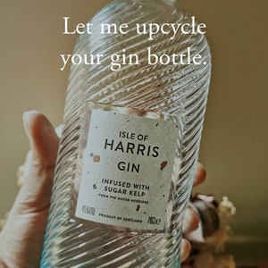 May include: A clear glass bottle of Isle of Harris Gin, infused with sugar kelp from the Outer Hebrides. The bottle has a blue and white swirl design and a wooden top.