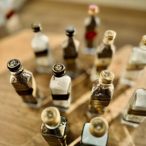 May include: A collection of small glass bottles with gold and black lids, arranged in a pattern on a wooden surface. The bottles are filled with different substances, such as spices or liquids.