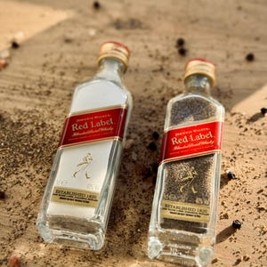 May include: Two clear glass bottles with red labels that say "Johnnie Walker Red Label Blended Scotch Whisky" and "Established 1820". The bottles are filled with black peppercorns and salt.