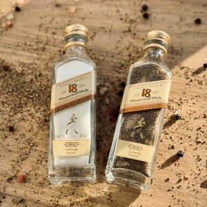 May include: Two small glass bottles filled with black peppercorns and salt. The bottles are labeled "Johnnie Walker 18" and have a gold cap. The bottles are sitting on a wooden surface.