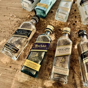 May include: A collection of seven glass bottles filled with sand and labeled with different varieties of Johnnie Walker Scotch whisky. The bottles are arranged on a wooden surface with scattered black peppercorns.