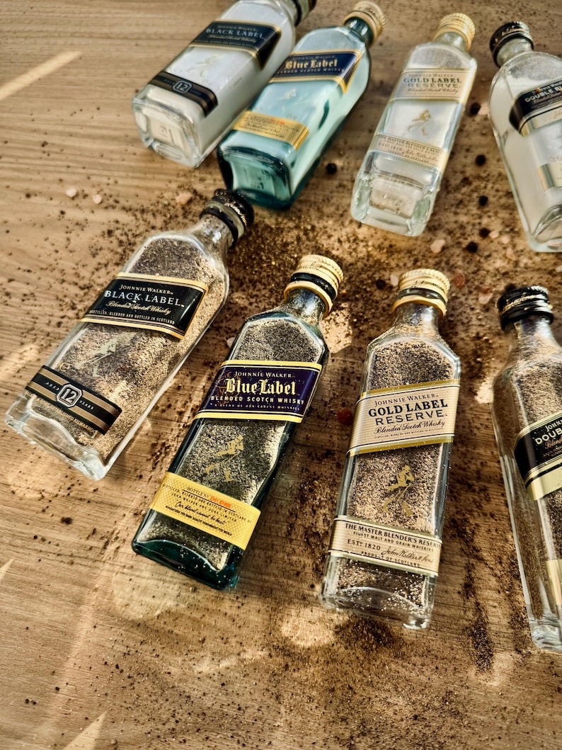 May include: A collection of seven glass bottles filled with sand and labeled with different varieties of Johnnie Walker Scotch whisky. The bottles are arranged on a wooden surface with scattered black peppercorns.