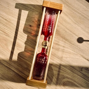 May include: A wooden hourglass-shaped stand with a clear glass bottle filled with a pink liquid. The bottle is labelled "Mermaid Pink Gin".