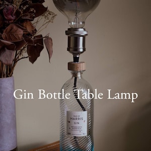 May include: A table lamp made from a clear glass gin bottle with a blue label that reads "Isle of Harris Gin" and a wooden top. The lamp has a clear glass light bulb with a black wire cord.