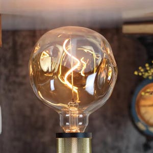 Op de afbeelding: Een heldere glazen gloeilamp met een decoratief, golvend filament. De lamp brandt en geeft een warm, geel licht. De lamp is bevestigd aan een zwarte en gouden metalen voet.